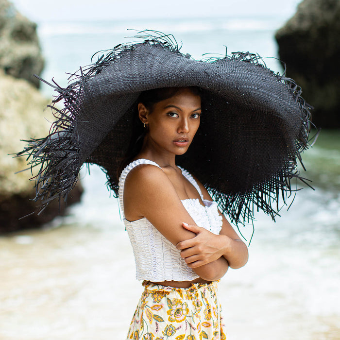 A woman is wearing BrunnaCo’s Amora Handwoven Frayed Oversized Straw Hat in black made by artisans in Bali. This tropical island hat is made for a perfect holiday vacation and coastal getaways. The photograph is taken by Faruq Adib’s team, Matarasa in Bingin Beach, Bali.