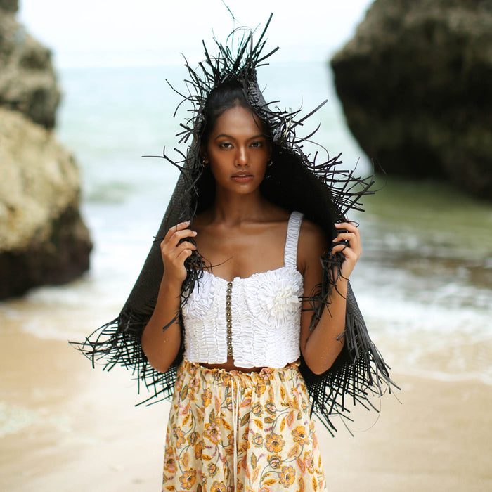 A woman is wearing BrunnaCo’s Amora Handwoven Frayed Oversized Straw Hat in black made by artisans in Bali. This tropical island hat is made for a perfect holiday vacation and coastal getaways. The photograph is taken by Faruq Adib’s team, Matarasa in Bingin Beach, Bali.