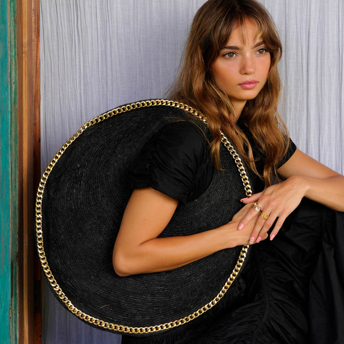 Inka Williams carries BrunnaCo's Black Fig Solo Bag, a handcrafted masterpiece made from sustainable jute fiber by skilled artisans in Bali. This elegant bag is adorned with a luxurious gold chain trim and offers ample space to hold all your beach essentials in style.