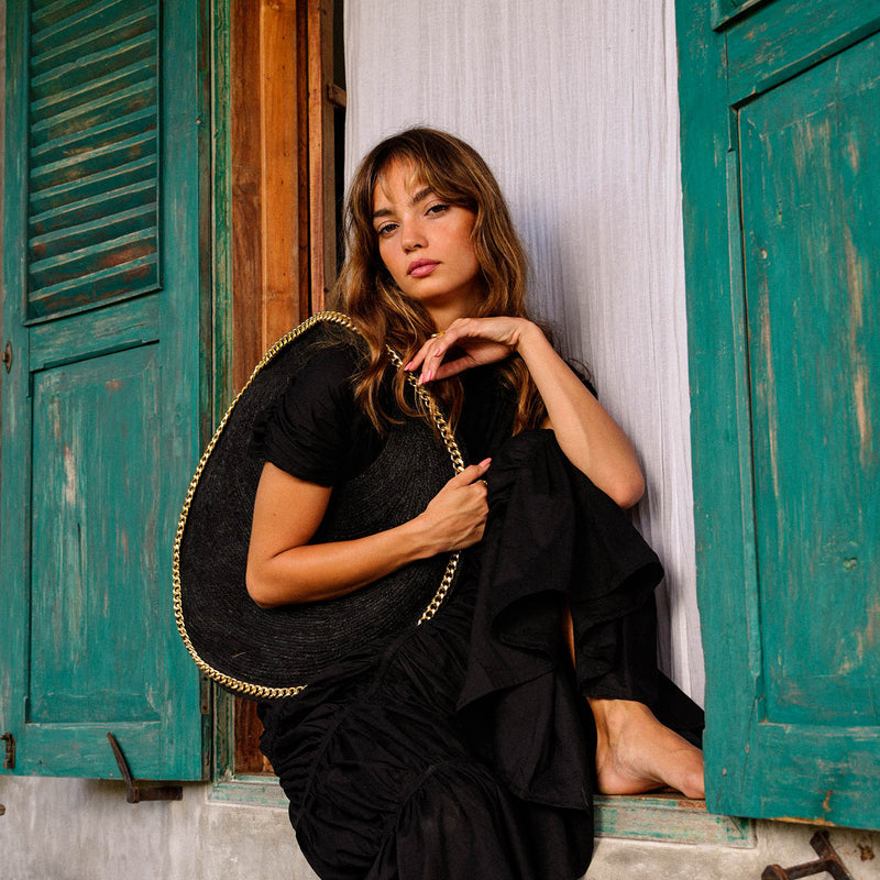 Inka Williams carries BrunnaCo's Black Fig Solo Bag, a handcrafted masterpiece made from sustainable jute fiber by skilled artisans in Bali. This elegant bag is adorned with a luxurious gold chain trim and offers ample space to hold all your beach essentials in style.