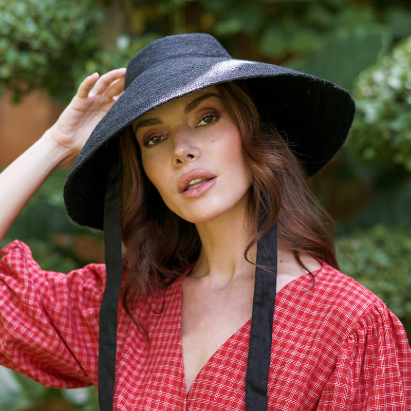 A woman is wearing BrunnaCo Reign Jute Straw hat for a summer stroll at the beach. The hat is sustainable, vegan, and made by artisans in Bali